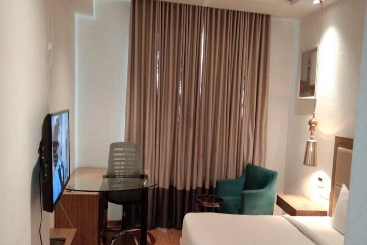 Une chambre d’hôtel comprenant un lit, un bureau avec une chaise, une télévision, une petite table et une fenêtre avec des rideaux.