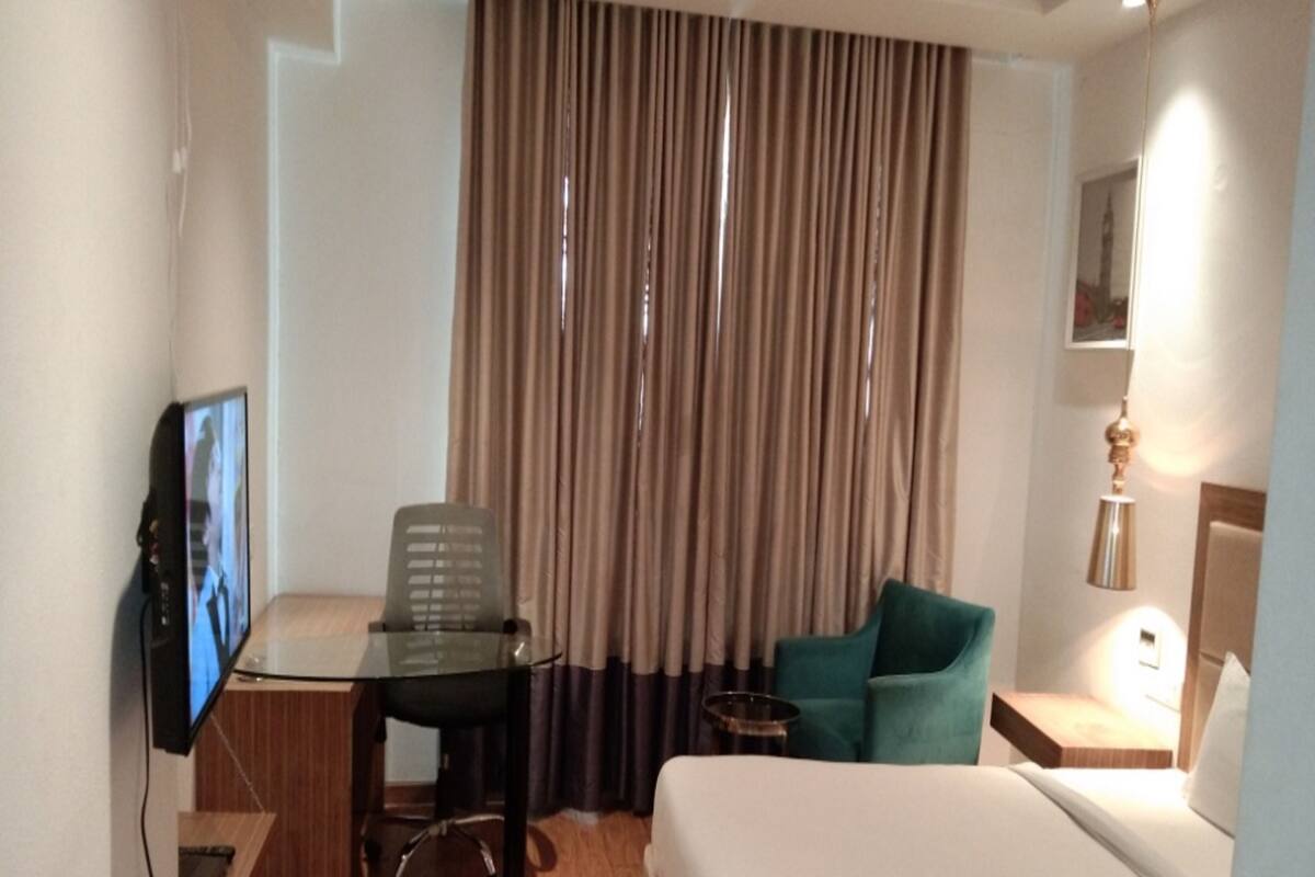 Une chambre d’hôtel comprenant un lit, un bureau avec une chaise, une télévision, une petite table et une fenêtre avec des rideaux.