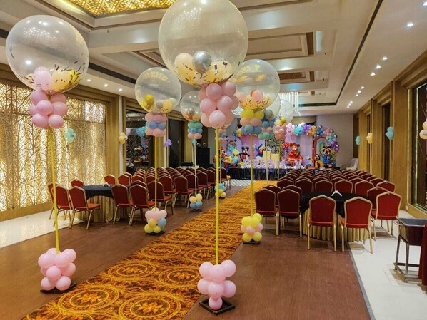 A banquet hall with rows of red chairs, a long table in the center, and balloons hanging from the ceiling. - Hotel Amora (Rajnandgaon)