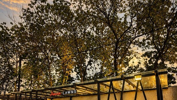 An outdoor garden area with a well-lit pathway, a seating area, and a wall with a name sign. - Hotel Amora (Rajnandgaon)