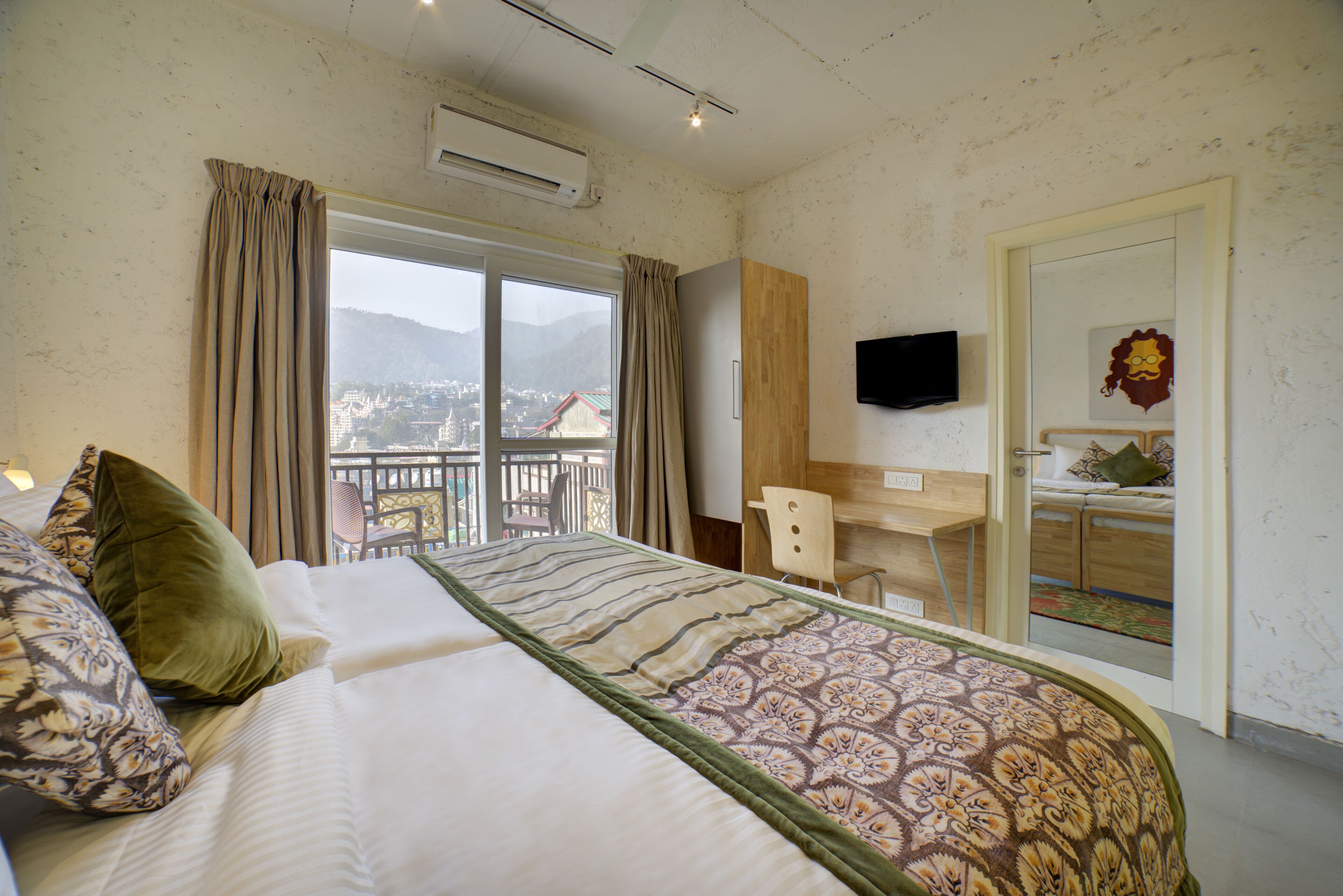 A hotel room with a bed, a desk, a chair, a television, and a balcony with a view of the city and mountains.