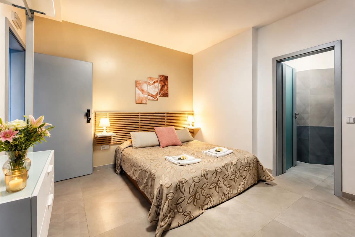 Een moderne hotelkamer met een bed, bedlampen, een vaas met bloemen en een deur naar de badkamer.