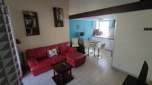 Una sala de estar con un sofá rojo, una mesa de centro y un televisor. Se ve una cocina al fondo con un microondas y una cafetera.