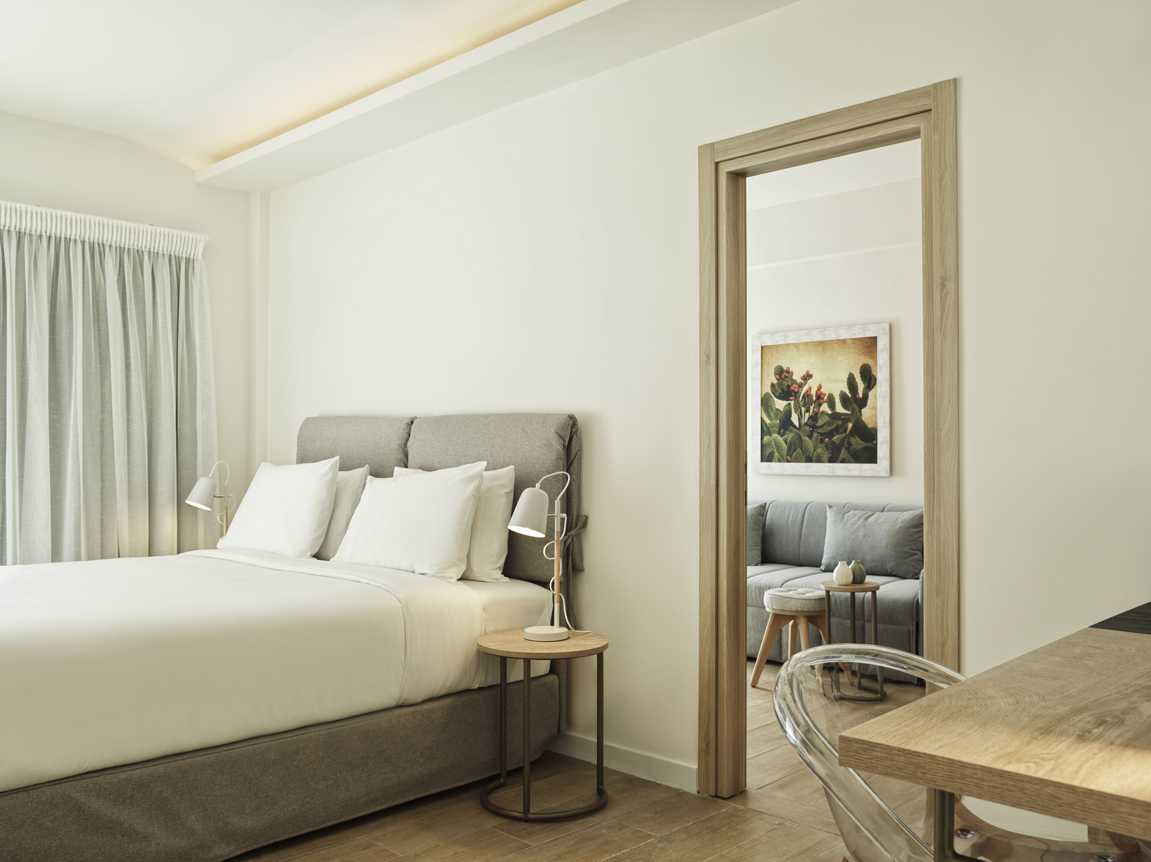 Una habitación de hotel moderna con una cama grande, una mesita de noche con lámpara, un rincón de descanso con sofá y una pequeña mesa, y un espejo grande en la pared.