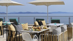 Un área de comedor al aire libre con mesas y sillas, sombrillas y vistas al mar y a las montañas.