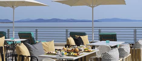 An outdoor dining area with tables and chairs, umbrellas, and a view of the sea and mountains.