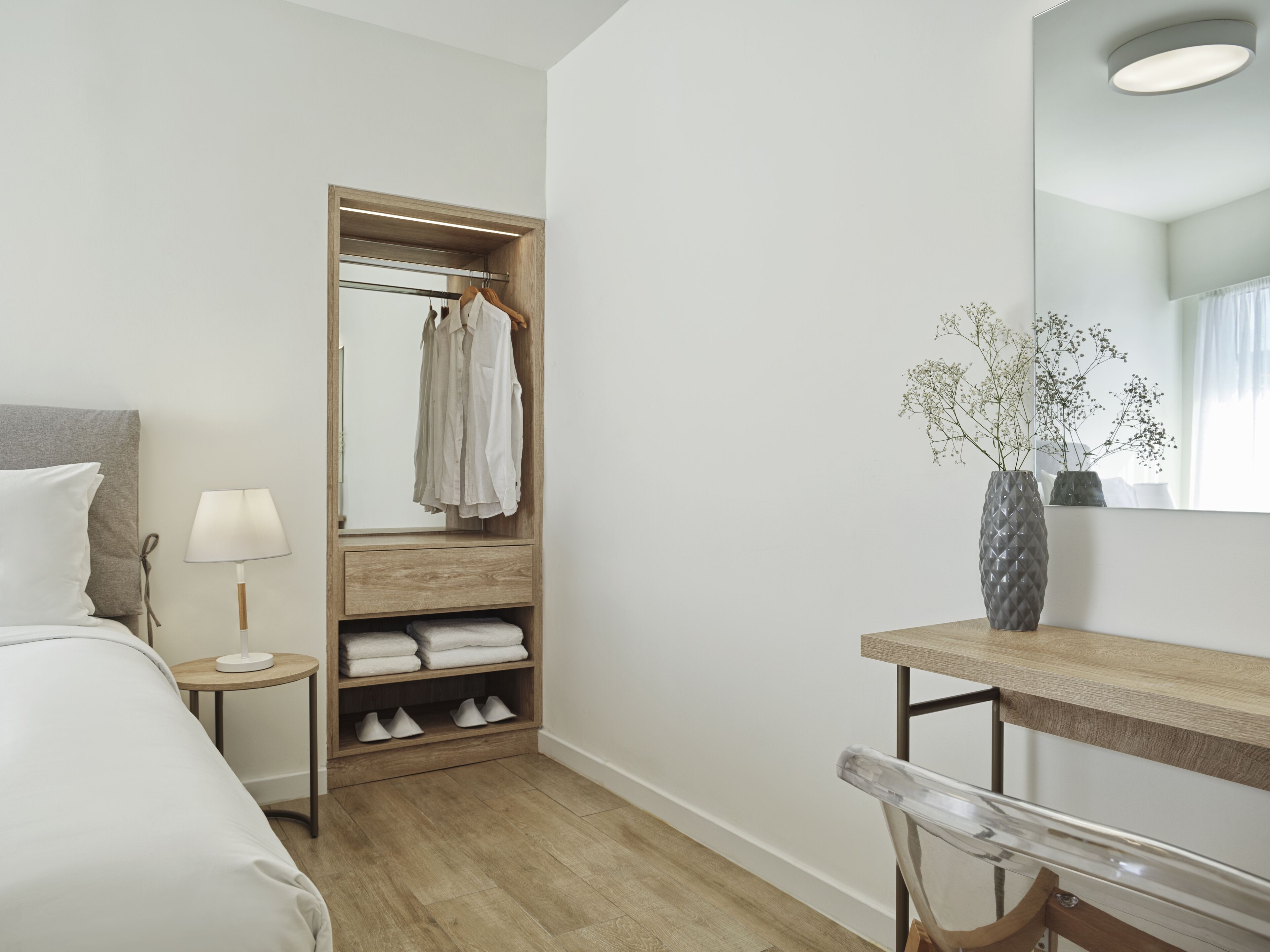 Hotelkamer met een bed, nachtkastje met lamp, houten garderobe met hangende kleding, spiegel, vaas met bloemen en bureau met stoel.