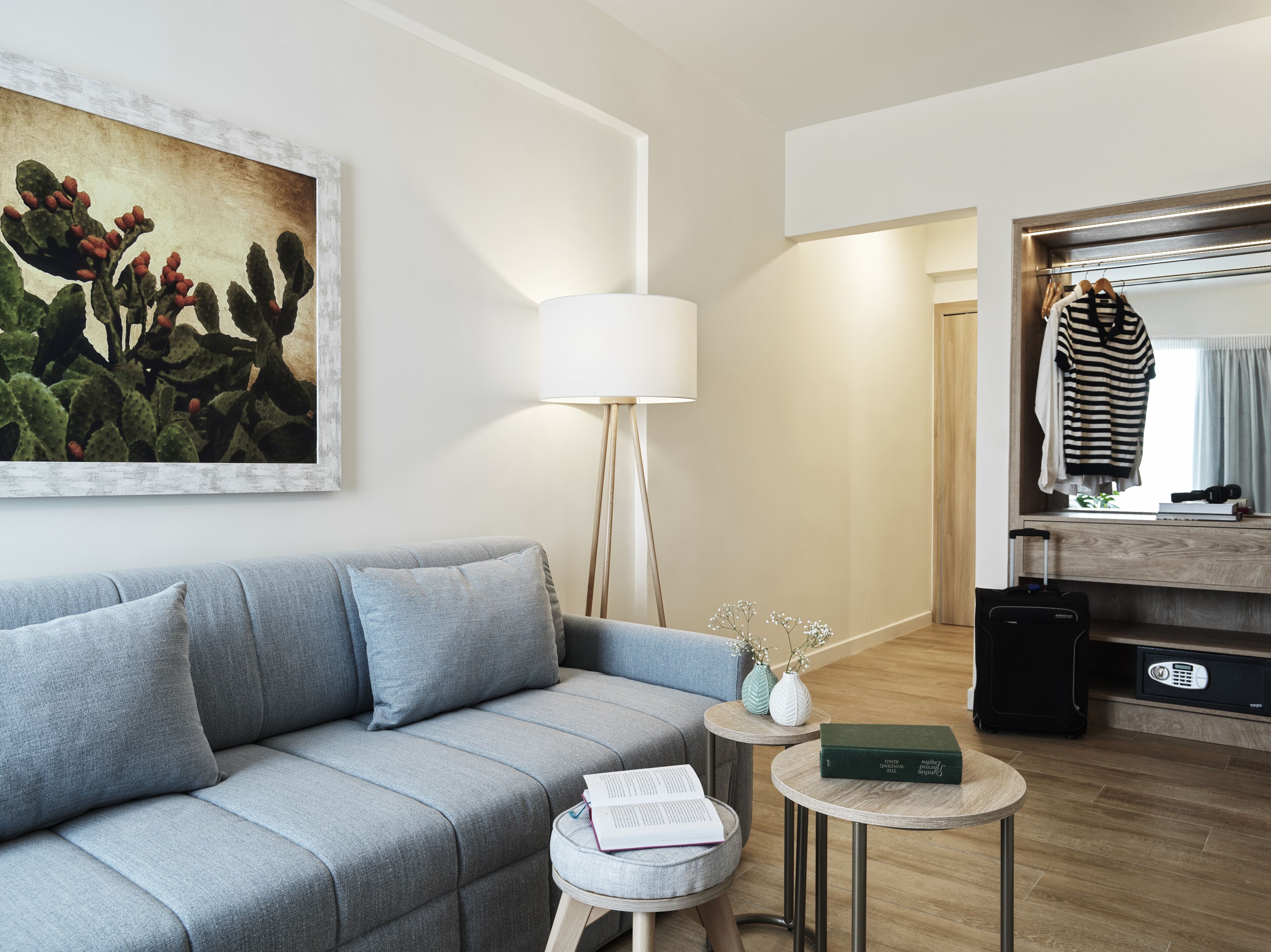 Un salon moderne avec un canapé gris, une lampe sur pied, une petite table ronde sur laquelle repose un livre, et un tableau représentant des cactus accroché au mur.