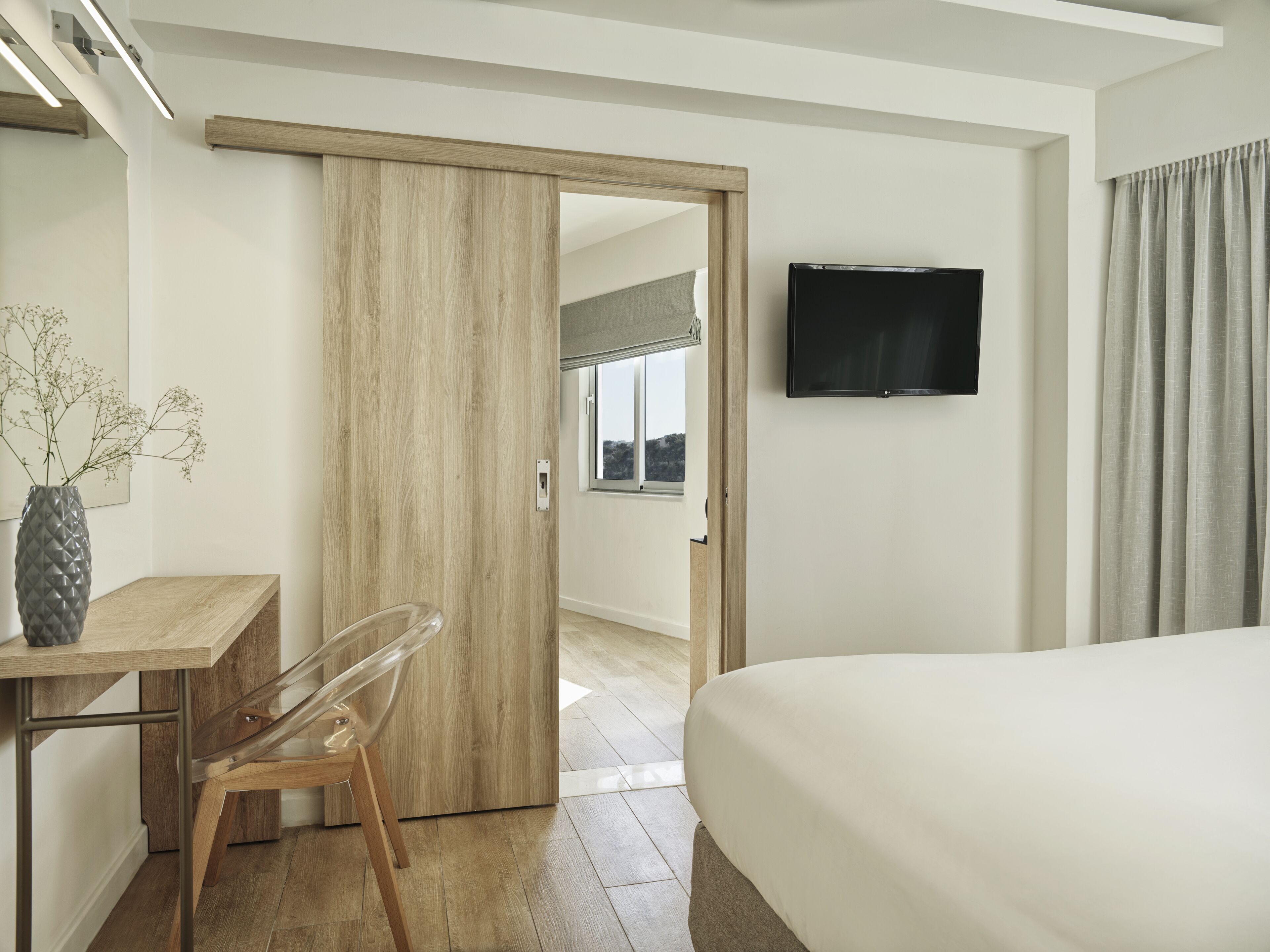 A hotel room with a bed, a desk with a chair, a TV mounted on the wall, and a sliding door leading to a balcony.