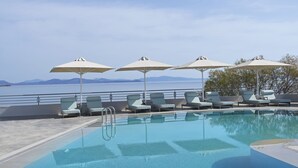 A pool area with lounge chairs and umbrellas, a clear view of the sea, and a mountainous horizon.