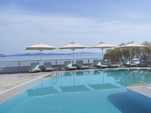 A pool area with lounge chairs and umbrellas, a clear view of the sea, and a mountainous horizon.