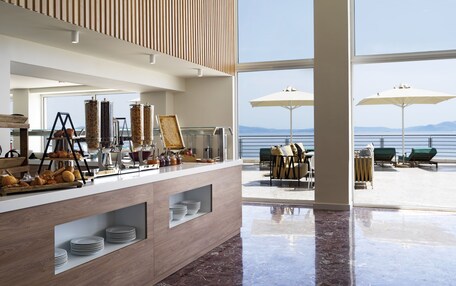 A modern breakfast bar with a view of the sea, outdoor seating with umbrellas, and a wooden ceiling.