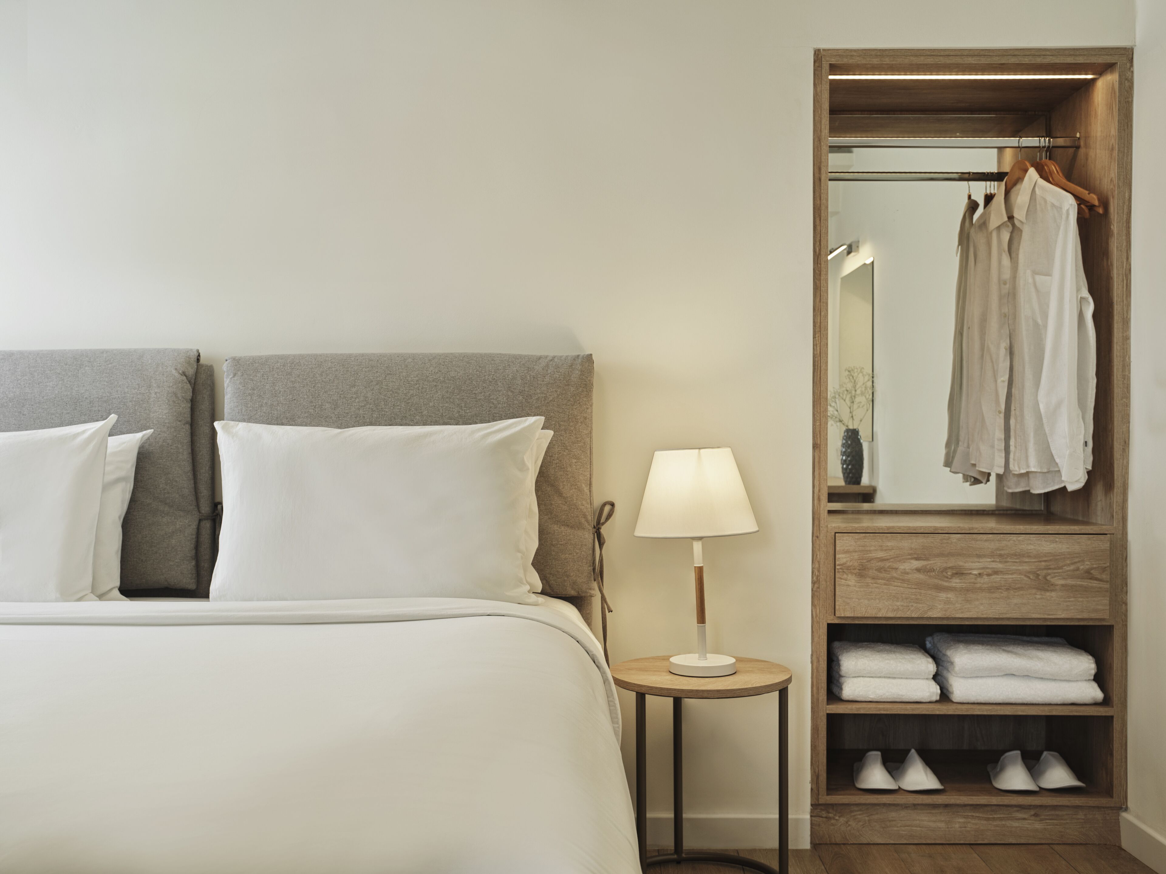 A neatly made bed with pillows, a bedside lamp, a wooden wardrobe with hanging clothes, and a shelf with folded towels.