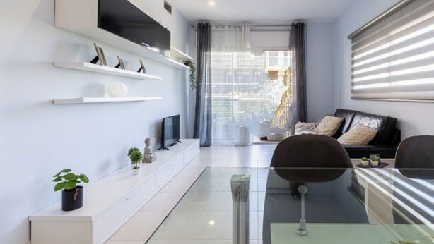 Ein modernes Wohnzimmer mit einem Glastisch, zwei schwarzen Stühlen, einer schwarzen Couch mit Kissen und einem Fernseher auf einem weißen Regal.