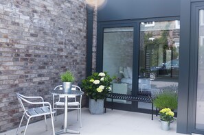 Un espace patio agrémenté d’un mur en briques, d’une porte vitrée, d’une table en métal avec des chaises et de plantes en pot.