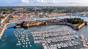 An aerial view of a harbor with numerous boats docked, a fortified structure, and a cityscape in the background.