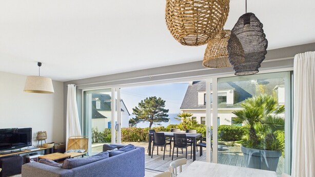 A modern living room with a sofa, TV, and dining table, large glass doors leading to a balcony with a view of the sea.