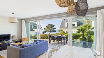 Een moderne woonkamer met een bank, televisie en eettafel, grote glazen deuren die uitkomen op een balkon met uitzicht op zee.