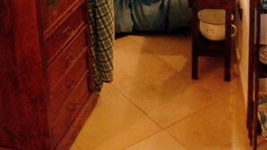 A bedroom with a bed, a wooden cabinet, a small table with a lamp, and a window with a checkered curtain.