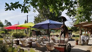 An outdoor seating area with wicker chairs and tables under umbrellas, a vintage cart, and a rustic wooden structure in the background.