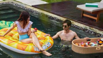 A pool with two people on inflatable floats, a wicker basket with food, and a wooden deck with a lounge chair.