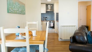 Un salon chaleureux avec une table à manger en bois, des chaises blanches et un canapé en cuir noir. On aperçoit au fond une cuisine avec des carreaux bleus et des meubles blancs.