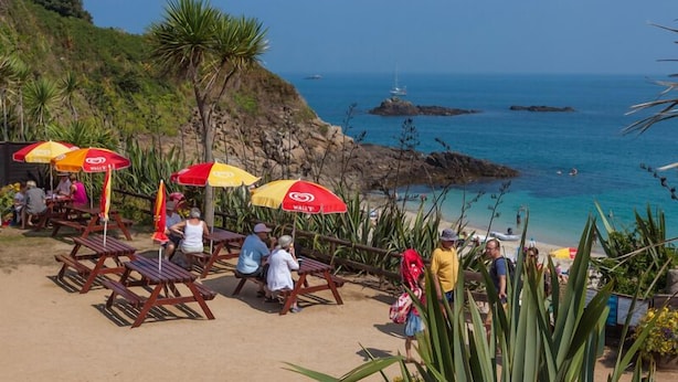 Une plage avec des chaises longues en bois et des parasols rouges, une côte rocheuse et l’océan en arrière-plan.