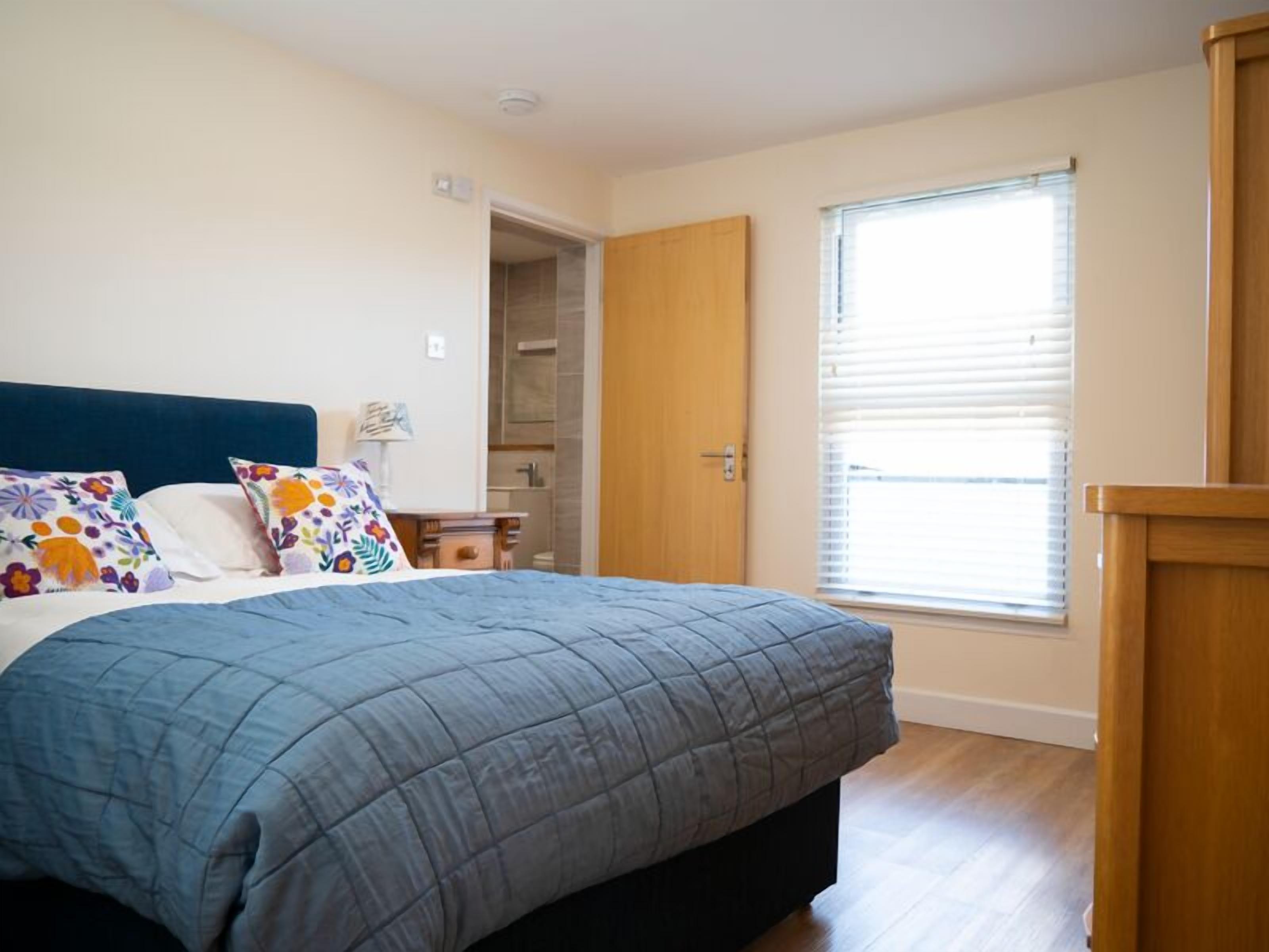 Un dormitorio con cama, mesita de noche, ventana con persianas y puerta que da a un baño.