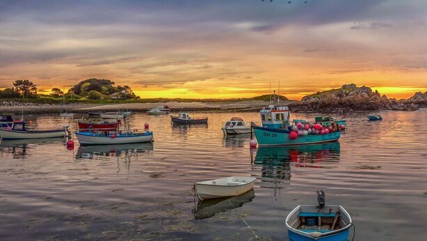 A harbor with several boats anchored, calm water, and a sunset in the background.