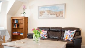 A living room with a brown leather sofa, a wooden bookshelf, a coffee table, and a painting of a coastal scene.