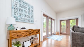 A spacious living room with hardwood flooring, a grey sofa, a wooden console table with decorative items, a blue patterned rug, and a large window with a view of trees. - Brand New 3 Bedroom, Pet Friendly Amazing Bay View Home (St George Island)