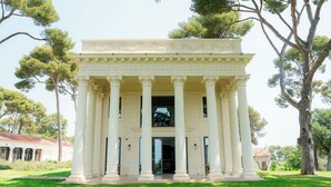 Un edificio neoclásico con una entrada central flanqueada por columnas, rodeado de un césped cuidado y árboles.