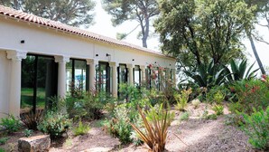 Un edificio con techo de tejas y ventanas amplias, rodeado por un jardín con diversas plantas y árboles.