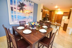 Un coin repas avec une table dressée pour quatre personnes, une cuisine en arrière-plan et un grand tableau aux motifs tropicaux accroché au mur.