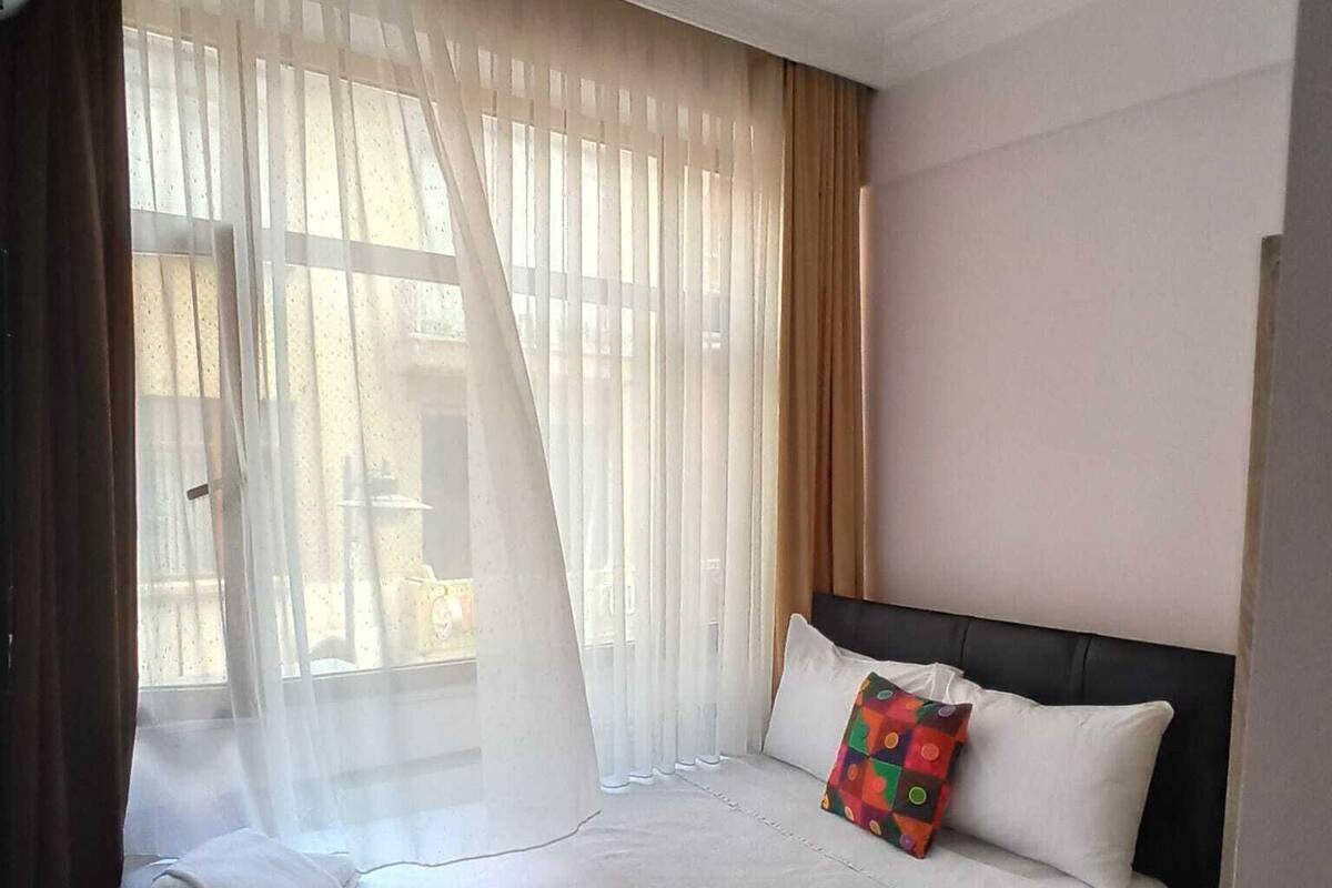 A hotel room with a bed, a nightstand, a window with sheer curtains, and a view of the outside.