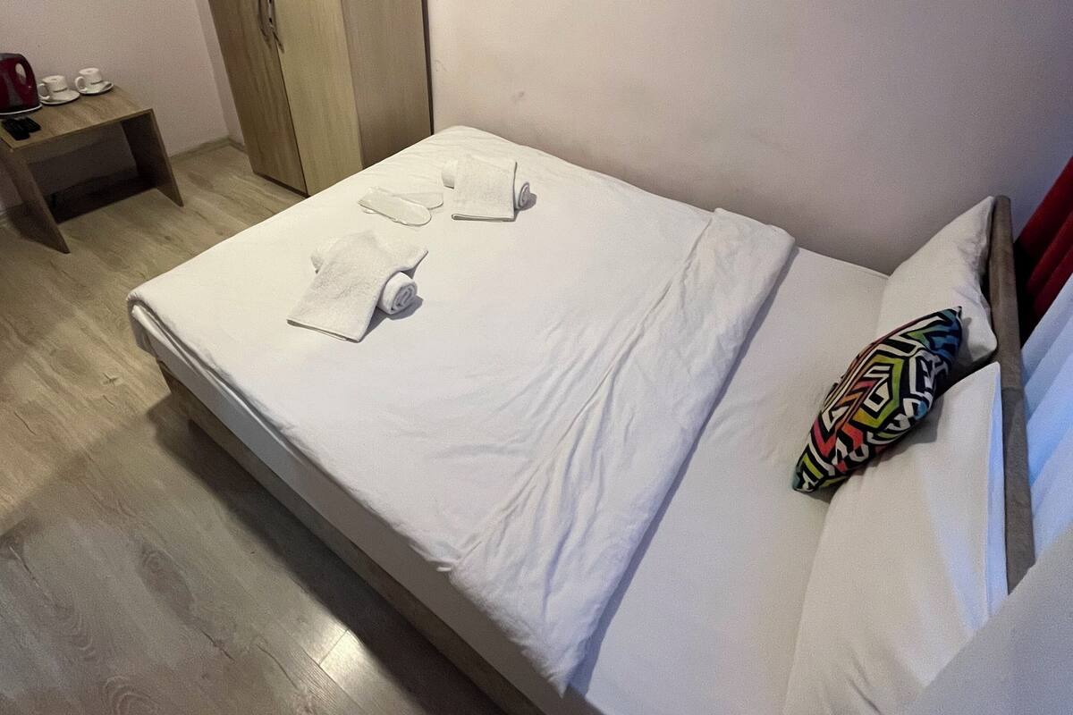 A neatly made bed with white linens and a colorful pillow, a wooden wardrobe, and a small bedside table with cups and a kettle.