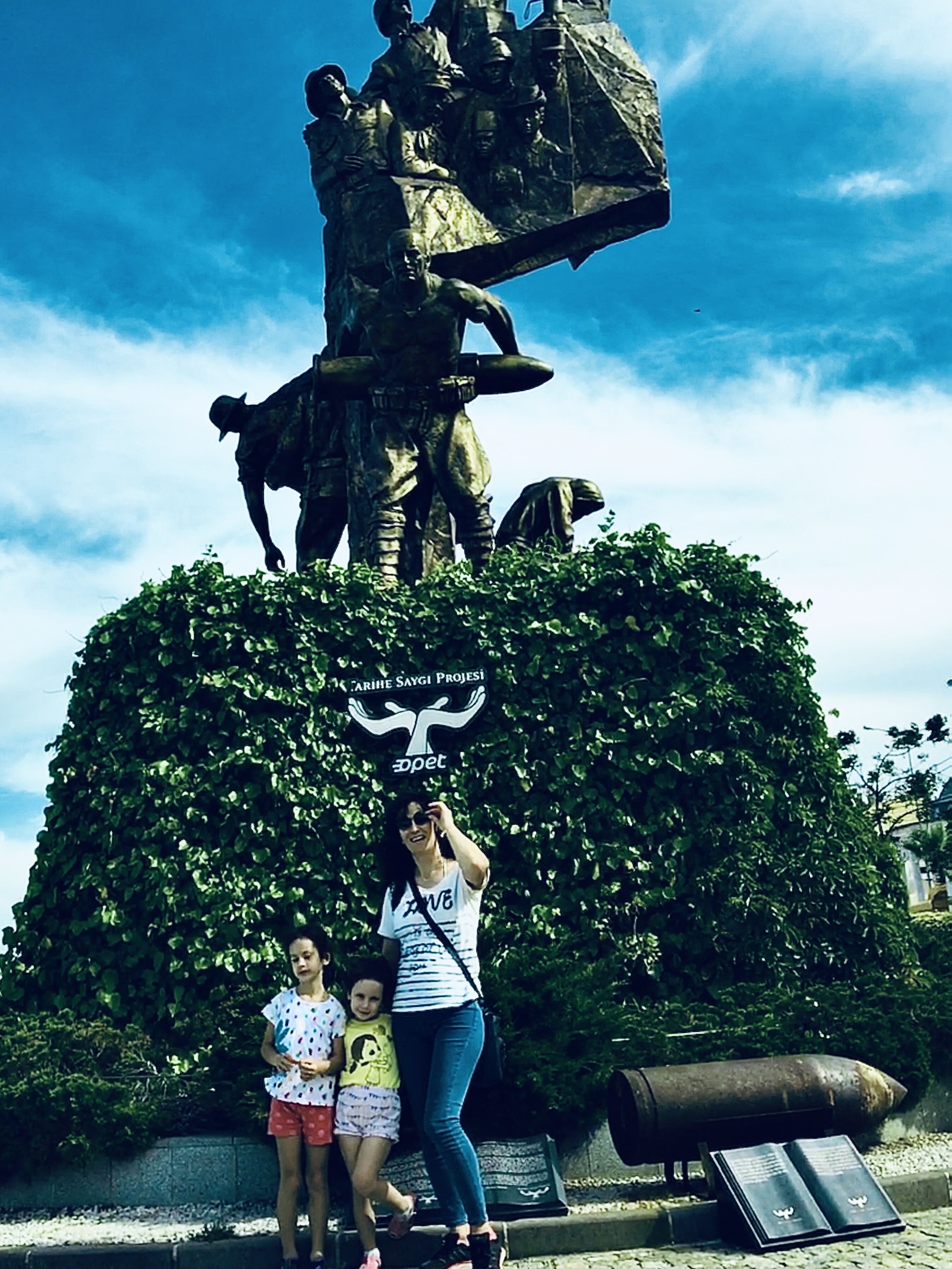 Una estatua de una figura histórica sobre un pedestal, rodeada de vegetación, con un cartel encima y placas informativas en la base.