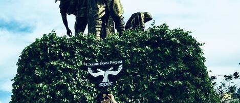 Una estatua de una figura histórica sobre un pedestal, rodeada de vegetación, con un cartel encima y placas informativas en la base.