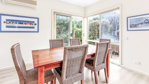 A dining area with a wooden table and wicker chairs, large windows, and framed artwork on the walls.