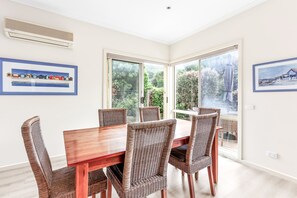 A dining area with a wooden table and wicker chairs, large windows, and framed artwork on the walls. - Edwards (Apollo Bay)