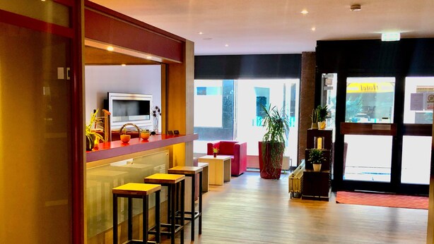 A modern bar area with stools, a counter, and a television. There are large windows and a plant in the center. - Montivo Hotel Köln-Bonn Airport (Troisdorf)