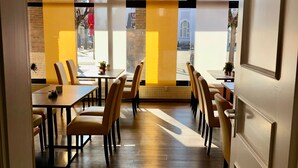 A modern dining area with wooden flooring, yellow accent walls, and large windows offering a view of the street. - Montivo Hotel Köln-Bonn Airport (Troisdorf)
