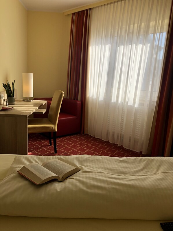 A hotel room with a bed, a desk with a lamp, a chair, a red sofa, and a large window with sheer curtains. - Montivo Hotel Köln-Bonn Airport (Troisdorf)