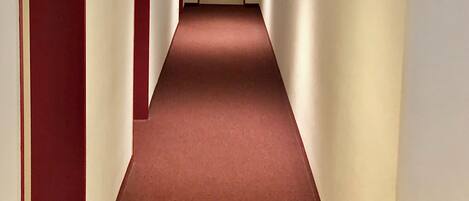 A narrow, carpeted hallway with white walls and red doors, leading to a closed door at the end.