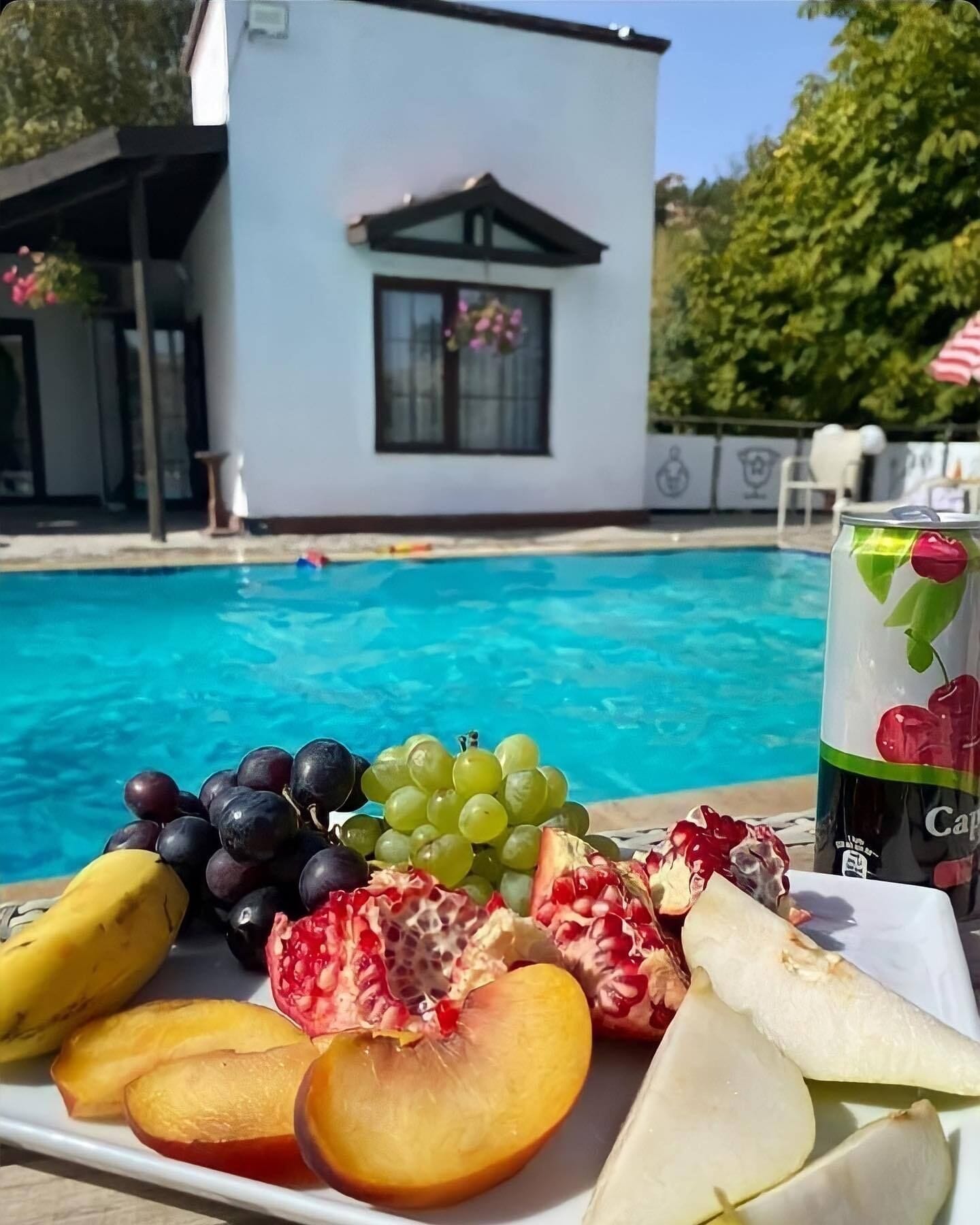 Une assiette de fruits frais, comprenant des raisins, des grenades, des pêches et des bananes, est placée au bord de la piscine, accompagnée d’une boisson décorée d’une cerise. On aperçoit en arrière-plan un bâtiment blanc doté d’une fenêtre et d’un balcon.