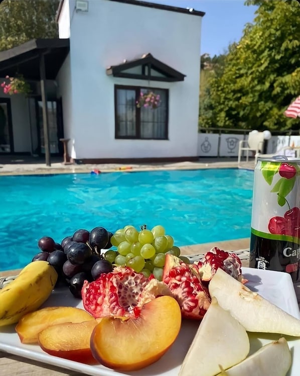 Una bandeja de frutas junto a la piscina con uvas, granada, melocotón y plátano, y una bebida con diseño de cereza, frente a un edificio blanco con ventana y balcón.