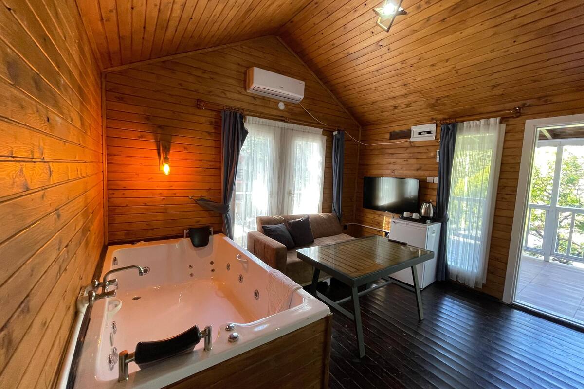 A wooden cabin interior with a large bathtub, a sofa, a TV, and a dining table.