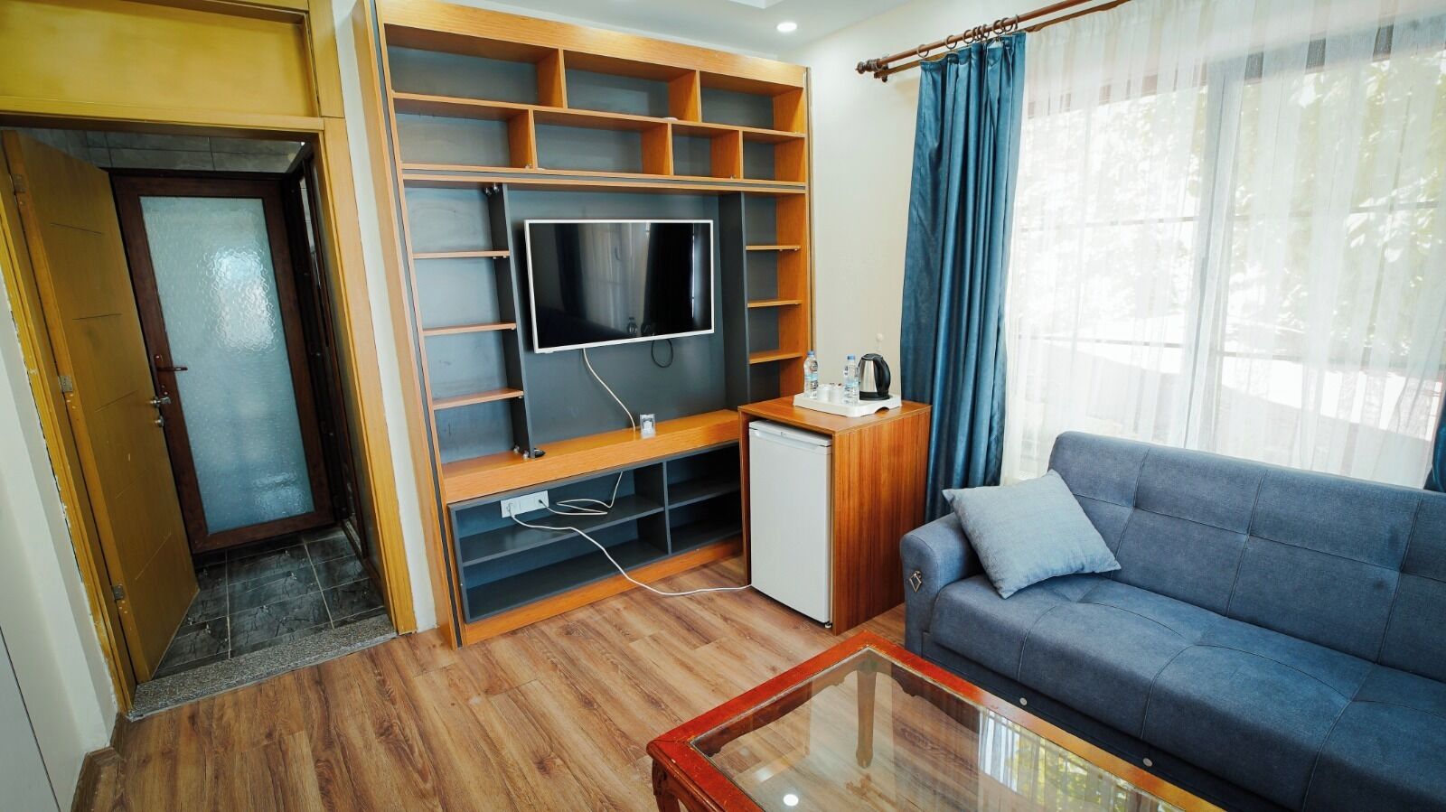 A living room with a sofa, a television, a small refrigerator, and a wooden shelving unit.