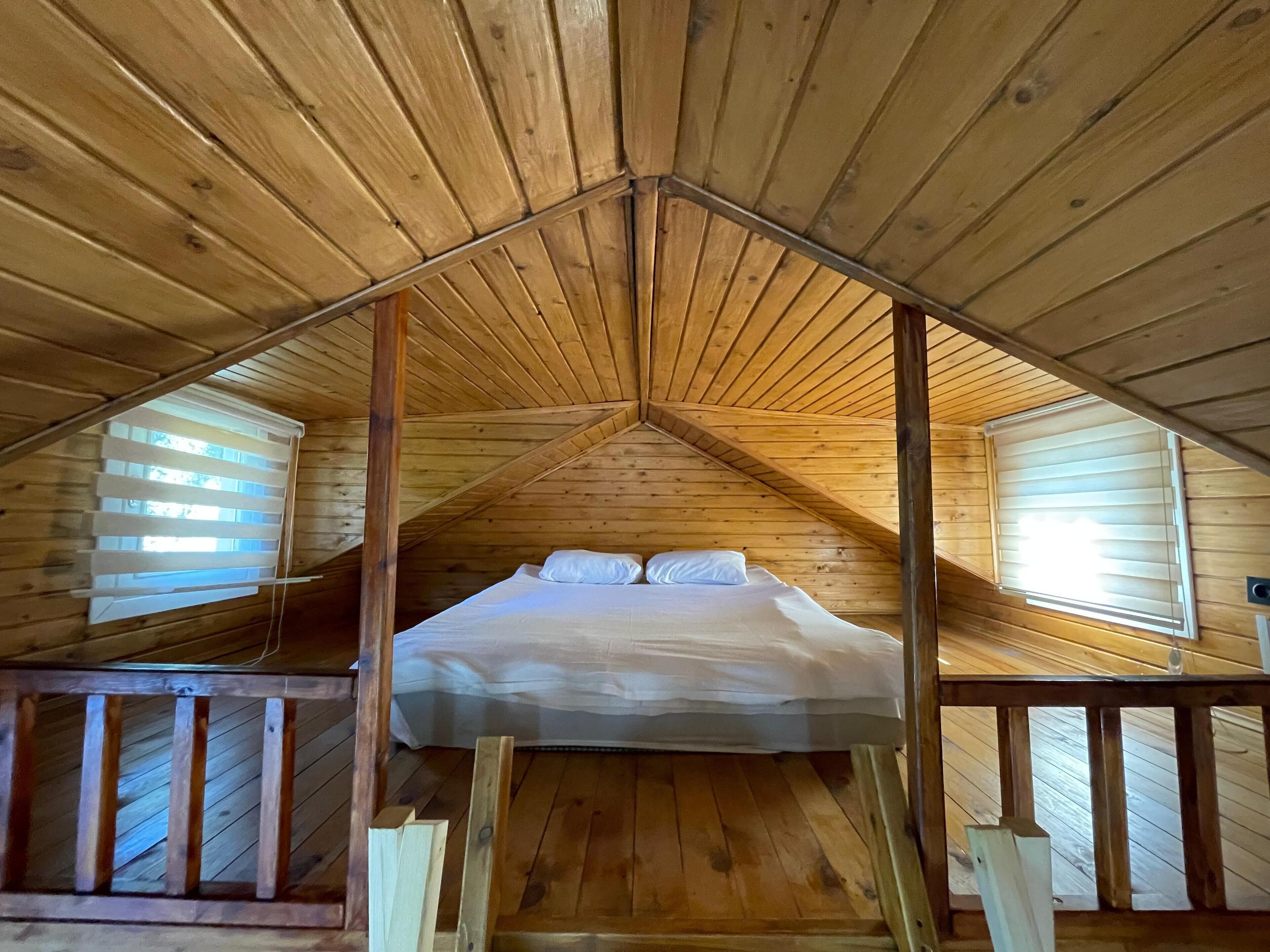 Una cabaña de madera con techo a dos aguas, una cama con ropa blanca y dos ventanas con persianas.