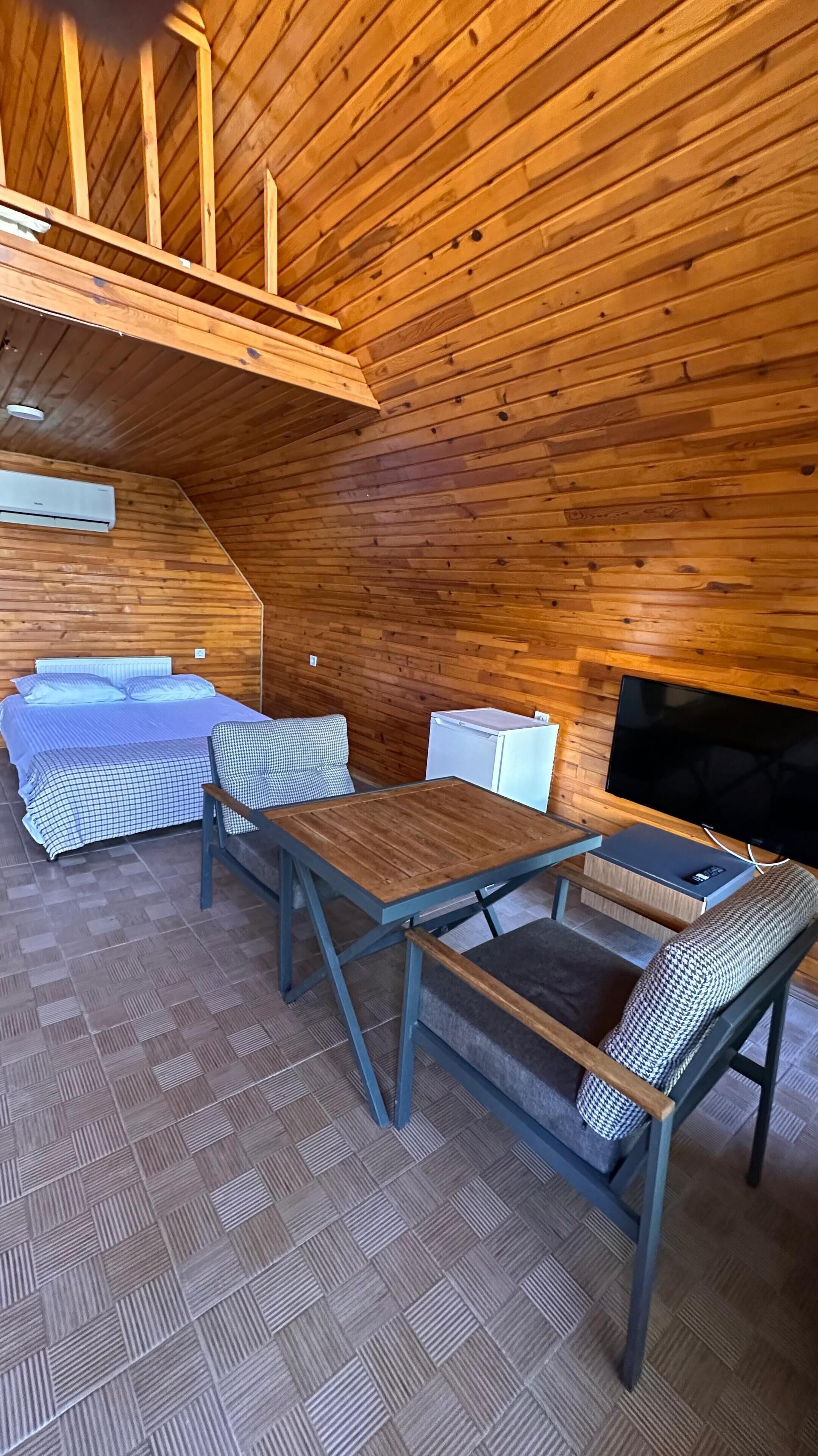 A cozy cabin interior with a wooden ceiling, a bed, a sofa, a small table, and a television.